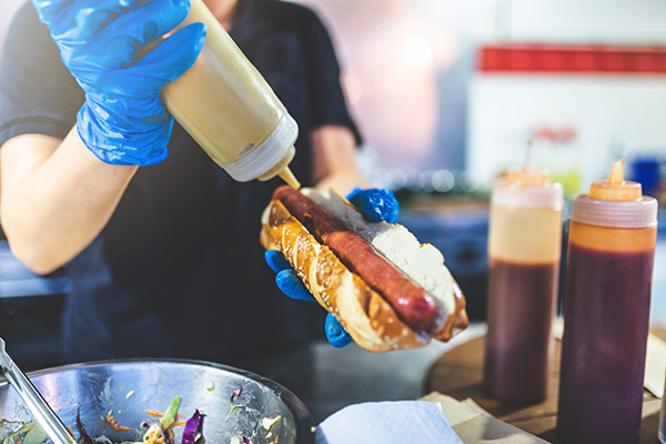 Close up of chef hands pouring mustard on fresh hot dog with grilled sausage.